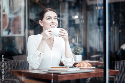Formal style. Beautiful businesswoman keeping smile on her face and holding cup in both hands while sitting in cafe