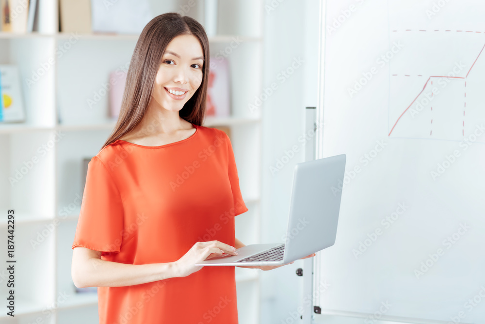 Data base. Merry glad pretty woman standing near white board while gazing at the camera and using laptop Data base. Merry glad pretty woman standing near white board while gazing at the camera and using laptop
