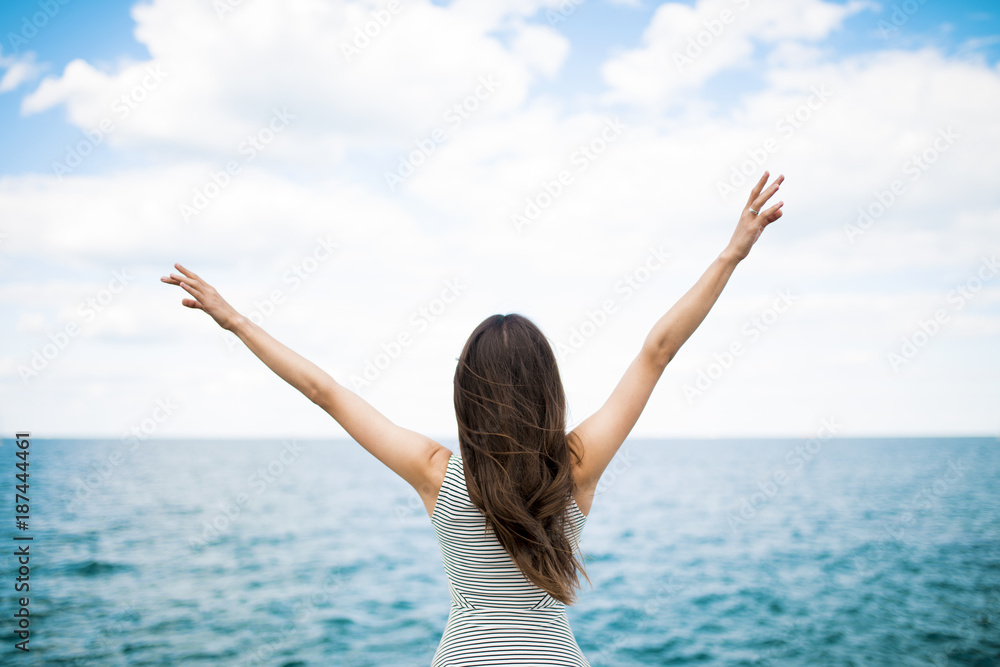 Rest and vacation concept. A young woman raises her hands to the sky against the background of the sea. Back view Rest and vacation concept. A young woman raises her hands to the sky against the background of the sea. Back view