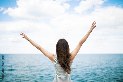 Rest and vacation concept. A young woman raises her hands to the sky against the background of the sea. Back view