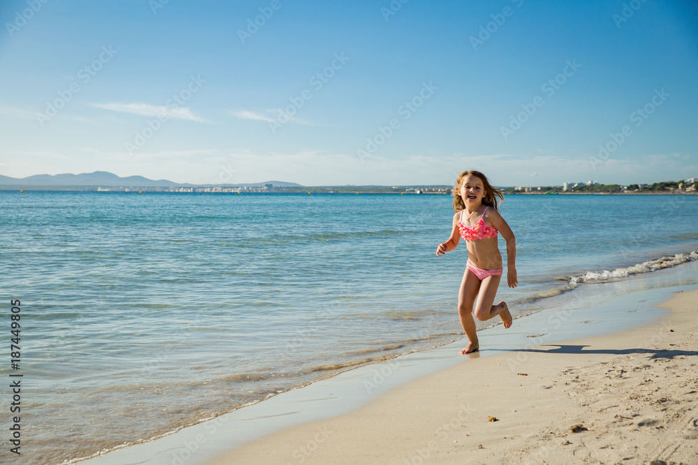Cute happy little girl running along the beach in swimming suit, jumping over waves. Beautiful summer sunny day, blue sea, turquoise water, picturesque landscape. Majorca, Spain