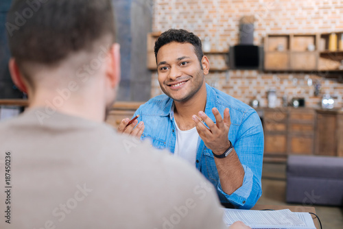 Pleasant smile. Positive happy emotional man sitting at the table and smiling cheerfully while looking at his smart pleasant colleagues