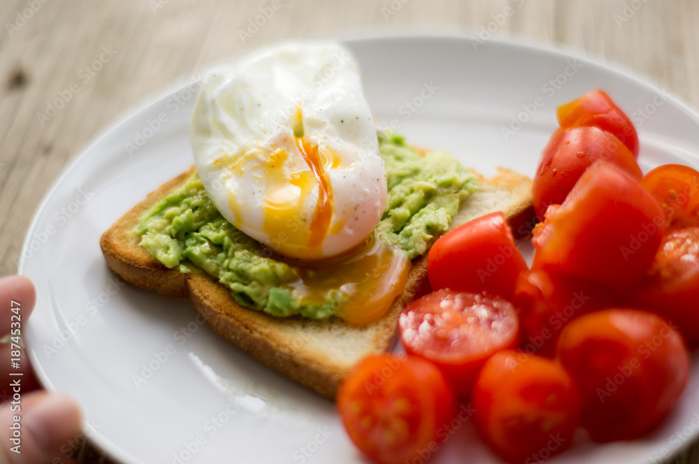 Perfect poached egg on a toast with smashed avocado and fresh tomatoes on a side on a white plate on the wooden table Perfect poached egg on a toast with smashed avocado and fresh tomatoes on a side on a white plate on the wooden table
