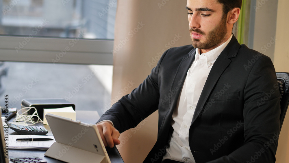 Young stylish man in suit using headphones and tablet while sitting at working desktop. Young stylish man in suit using headphones and tablet while sitting at working desktop.