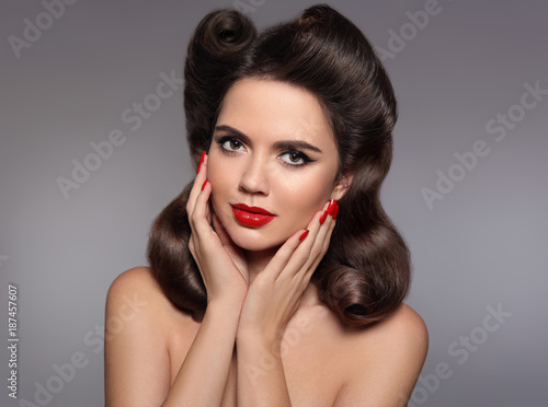 Pin up hairstyle. Beautiful 50s girl holding her cheeks with red lips makeup and manicured nails looking at camera. Expressive facial expressions. Beauty fashion studio photo.
