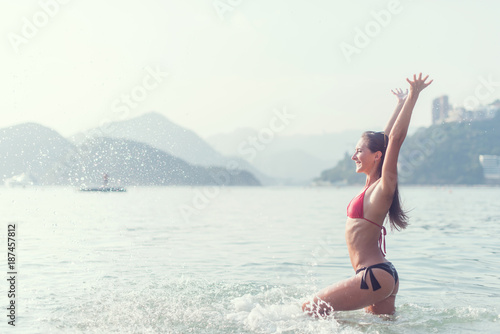 Happy young woman excited about holidays splashing water by raising her hands up standing in the sea. Cheerful girl tourist in bikini enjoying bright sunshine. © undrey