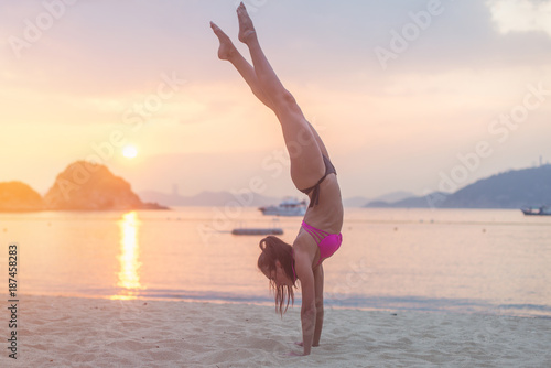 Young fitness woman doing handstand exercise on beach at sunrise. Sporty girl in bikini practicing yoga seashore.