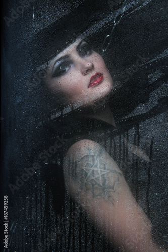 portrait of a beautiful angry brunette with a tattoo of a pentagram behind wet glass after rain