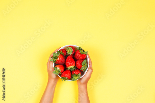 Kid holds in hand bowl with fresh strawberries on yellow background