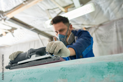 Portrait of worker wearing respirator repairing boat in yacht workshop using electric polishing tool, focus on foreground