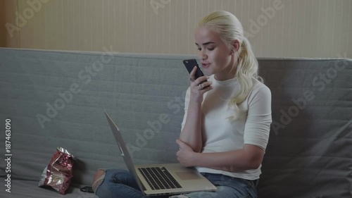 Girl using a smart phone voice recognition on line sitting on a sofa in the living room at home with a warm light blonde model student photo