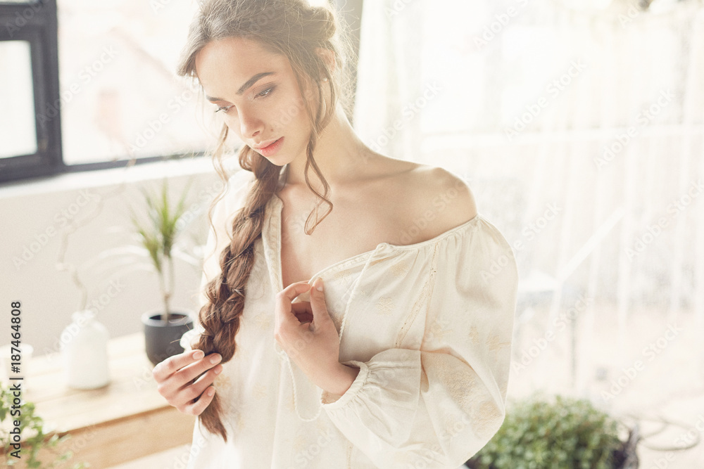 Sunny portrait of beautiful sexy and young woman with dark braid wearing simple light dress in studio with big windows, plants, bath and straw chair Sunny portrait of beautiful sexy and young woman with dark braid wearing simple light dress in studio with big windows, plants, bath and straw chair