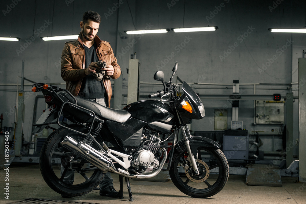 Fototapeta premium Młody człowiek stoi blisko motocyklu w garażu w skórzanej kurtce