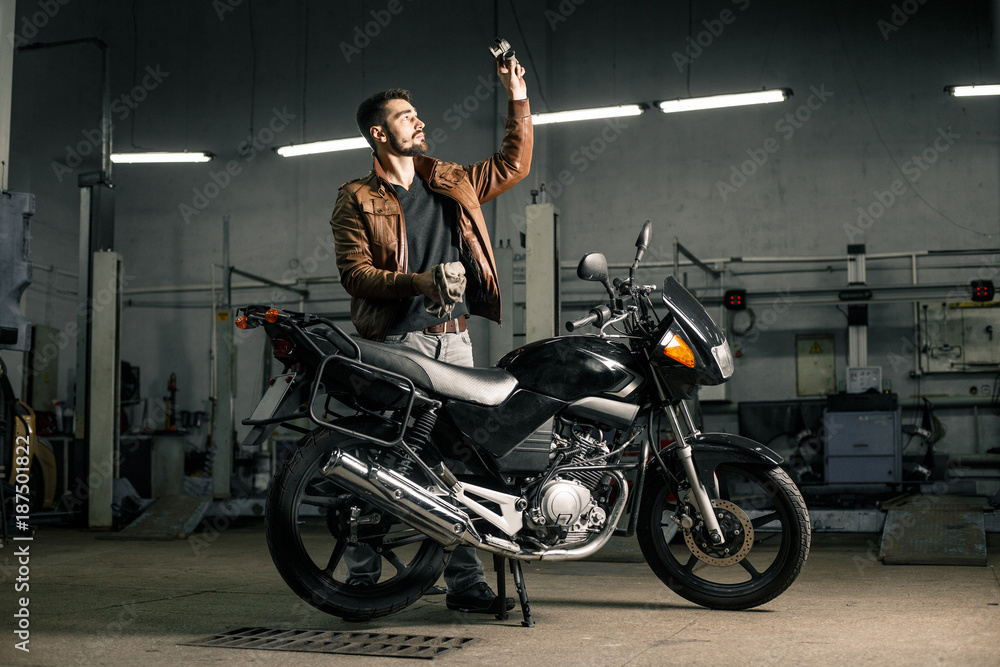 Young man in brown leather jacket standing near motorcycle in garage