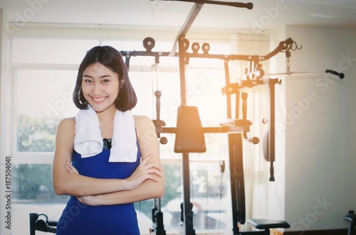 young happy smiling asian woman slim body with towel crossing arms in fitness gym with machine in background, bodybuilder, healthy lifestyle, exercise, workout and sport training concept