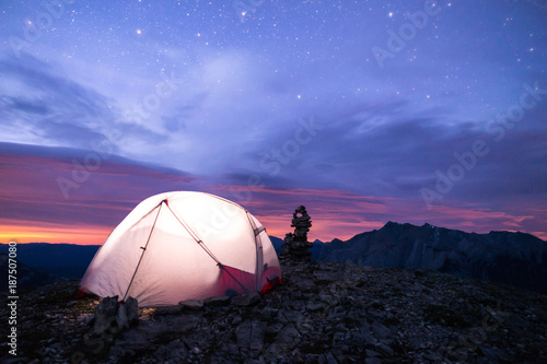 Lit up tent under the stars on a mountain. There is light pollution that looks like a sunset or sunrise with a layer of clouds in between the starry night sky. There is an inukshuk beside the tent.