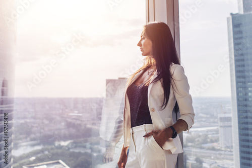 Portrait of elegant business lady wearing white formal suit standing near window looking at cityscape