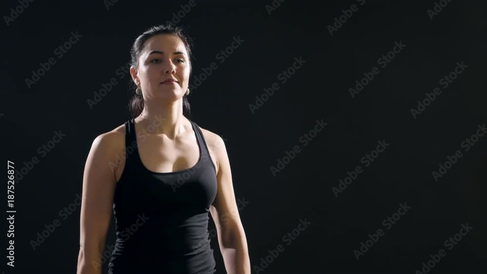 An impressive view of a young lady who stands and raises dumbbells in her hands up by turns before her breast in an active way in a black studio. An impressive view of a young lady who stands and raises dumbbells in her hands up by turns before her breast in an active way in a black studio.