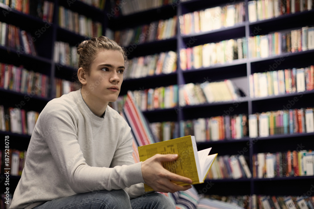 Young Caucasian guy wearing gray sweater spending day at bookstore searching for book on art, holding yellow one in his hands, having uncertain look. People, reading, leisure and knowledge concept Young Caucasian guy wearing gray sweater spending day at bookstore searching for book on art, holding yellow one in his hands, having uncertain look. People, reading, leisure and knowledge concept