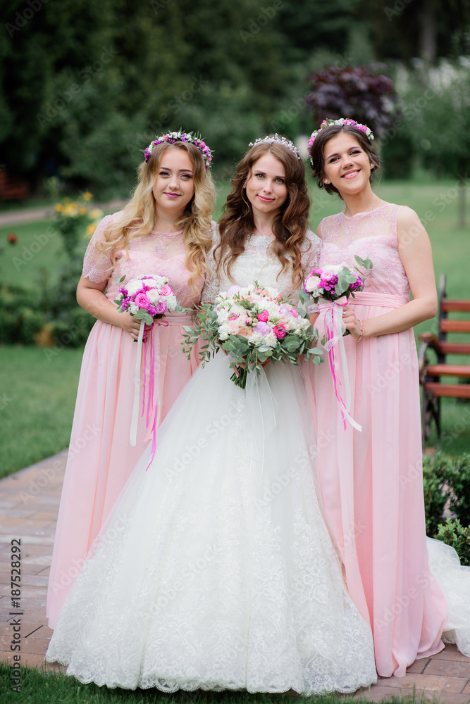 Stunning young bride and bridesmaids in pink dresses stand with wedding bouquets in the park