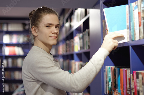Picture of handsome student guy with hair gathered in knot looking at camera with cute smile, picking book from bookshelf in library or bookstore. Literature, learning and knowledge concept