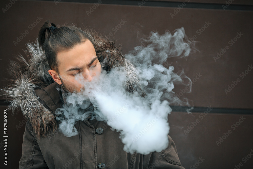 Vape man. Portrait of a handsome young white guy with modern haircut vaping an electronic cigarette opposite the futuristic urban background. Lifestyle.