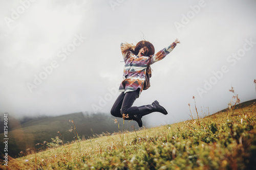 stylish traveler girl in hat with backpack jumping in mountains. happy hipster woman having fun on top of mountain. space for text. wanderlust and travel concept. atmospheric moment