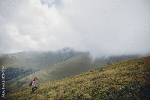Fototapeta Naklejka Na Ścianę i Meble -  wanderlust and travel concept. girl traveler in hat with backpack walking in clouds in mountains. stylish hipster woman exploring on top of mountain. space for text. atmospheric moment.