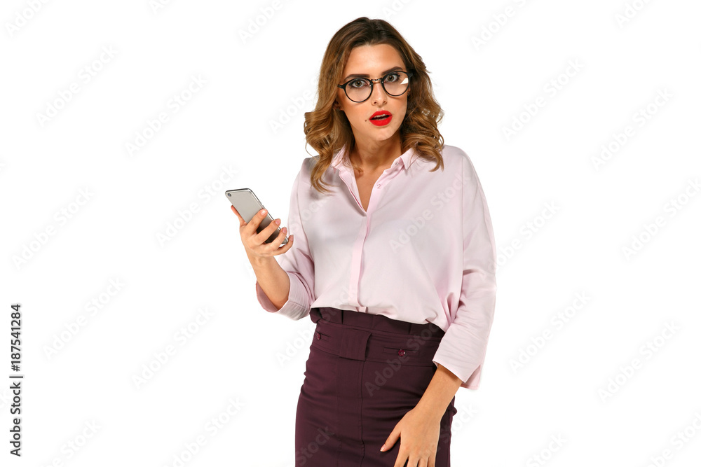 Pretty surprised, indignant business woman holding mobile phone, dressed up in blouse and skirt, in eyeglasses, with red lips, wavy hair, on white background