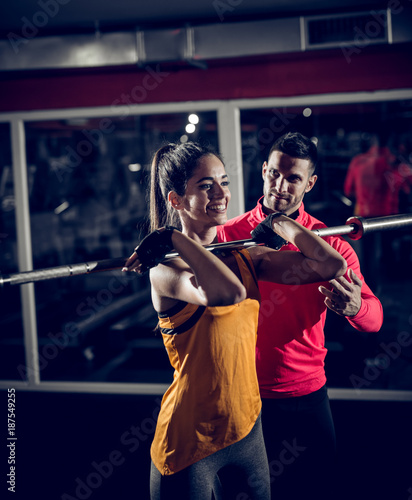 Young smiling healthy sporty strong active shape girl holding barbell without weights on the shoulder in front while helpful personal trainer standing next to her.