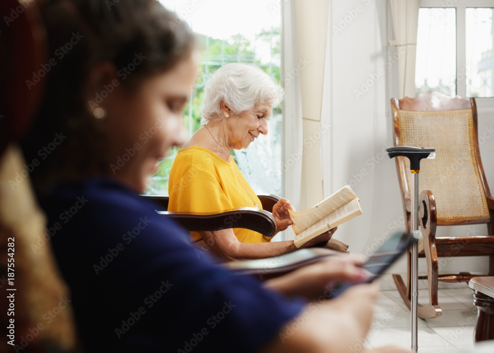 Granddaughter Using Mobile Phone And Grandma Reading Book Granddaughter Using Mobile Phone And Grandma Reading Book