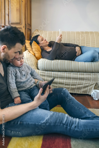 Young father looking at the tablet with his little son while the pregnant mother looks the mobile. Focus on pregnant woman in background