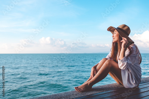 Portrait image of a happy beautiful asian woman on white dress sitting on wooden balcony by the sea with clear blue sky background