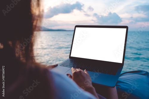 Mockup image of a woman using laptop with blank white desktop screen while sitting by the sea with blue sky background