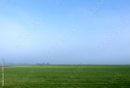 Wide view of a misty green farming field with a barn in the distance and a blue sky.