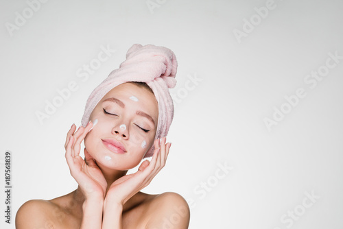 a cute young girl with a towel on her head puts a nourishing cream on her face, her eyes are closed, she enjoys