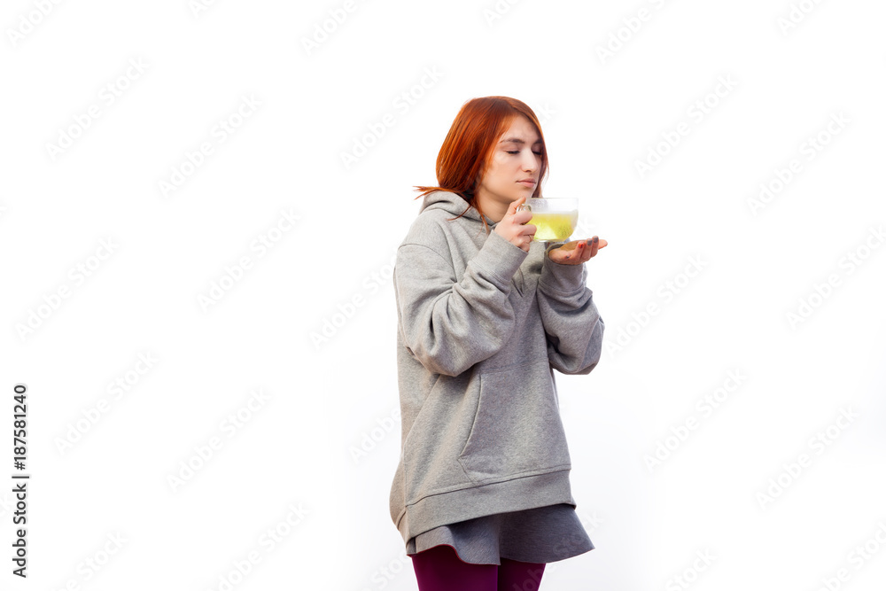 Young red-haired woman in sports sweatshirt gets sick and drinks from large transparent mug antiviral medicine with vitamin with thermometer on white isolated background Young red-haired woman in sports sweatshirt gets sick and drinks from large transparent mug antiviral medicine with vitamin with thermometer on white isolated background