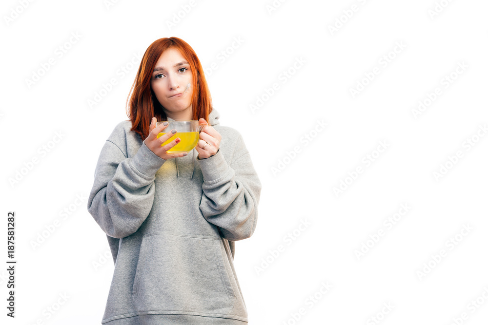 Young red-haired woman in sports sweatshirt gets sick, drinks from large transparent mug antiviral medicine and measures temperature by thermometer on white isolated background Young red-haired woman in sports sweatshirt gets sick, drinks from large transparent mug antiviral medicine and measures temperature by thermometer on white isolated background