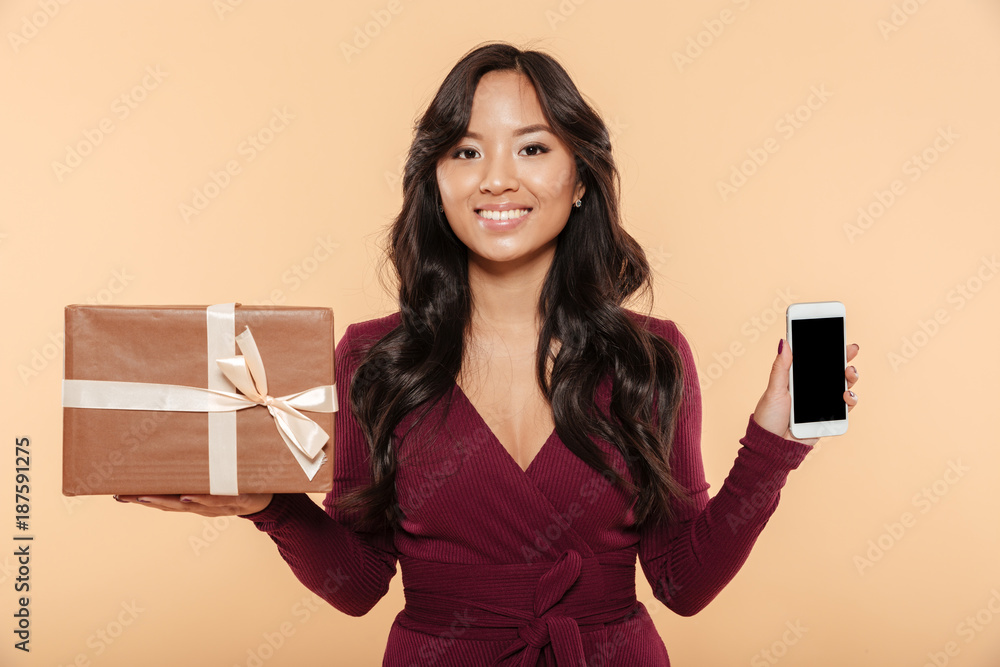 Asian smiling woman in maroon dress demonstrating present box with smartphone as gift being isolated over peach background Asian smiling woman in maroon dress demonstrating present box with smartphone as gift being isolated over peach background