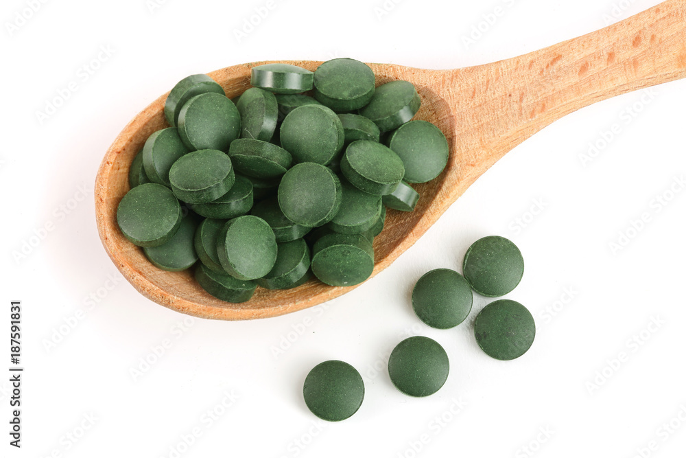 heap of Spirulina tablets algae nutritional supplement in wooden spoon isolated on white background close up top view