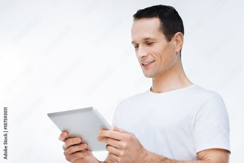 Digital device. Joyful positive young man holding a tablet and looking at its screen while using modern technology Digital device. Joyful positive young man holding a tablet and looking at its screen while using modern technology