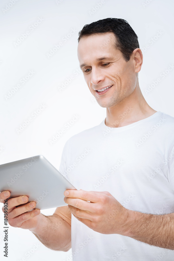 Modern device. Happy cheerful nice man smiling and looking at the tablet screen while surfing on the Internet Modern device. Happy cheerful nice man smiling and looking at the tablet screen while surfing on the Internet