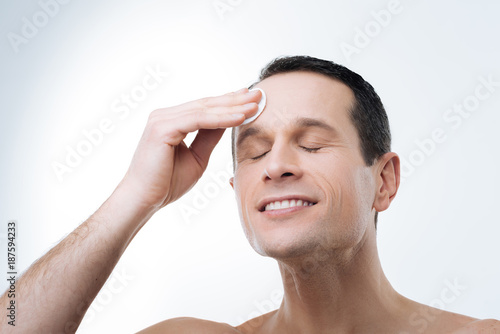 Pleasant procedure. Nice delighted positive man closing his eyes and holding a cotton pad while cleaning his forehead