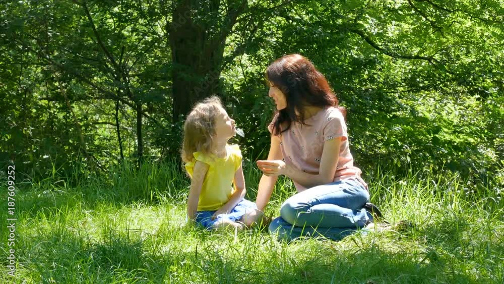 Happy Mother and Little Daughter Playing at the Park Having Picnic Outdoors Sitting on the Green Grass. Young Pretty Brunette is Talking to a Cute Girl. Happy Mother and Little Daughter Playing at the Park Having Picnic Outdoors Sitting on the Green Grass. Young Pretty Brunette is Talking to a Cute Girl.