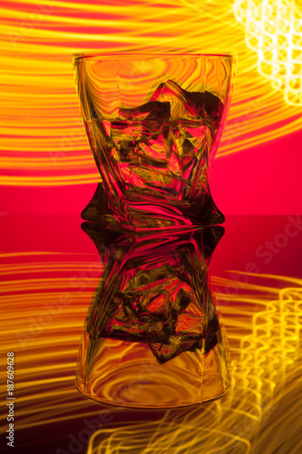 Cocktail whiskey a glass with pieces ice of party reflection a concept of hourglasses yellow light effects on red background.