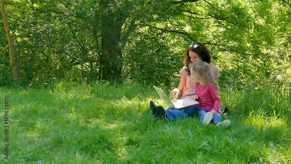Family Spend Time Together Outdoors Using White Laptop. Two Girls are Playing Video Games Sitting on the Green Grass in the City Park. Family Spend Time Together Outdoors Using White Laptop. Two Girls are Playing Video Games Sitting on the Green Grass in the City Park.