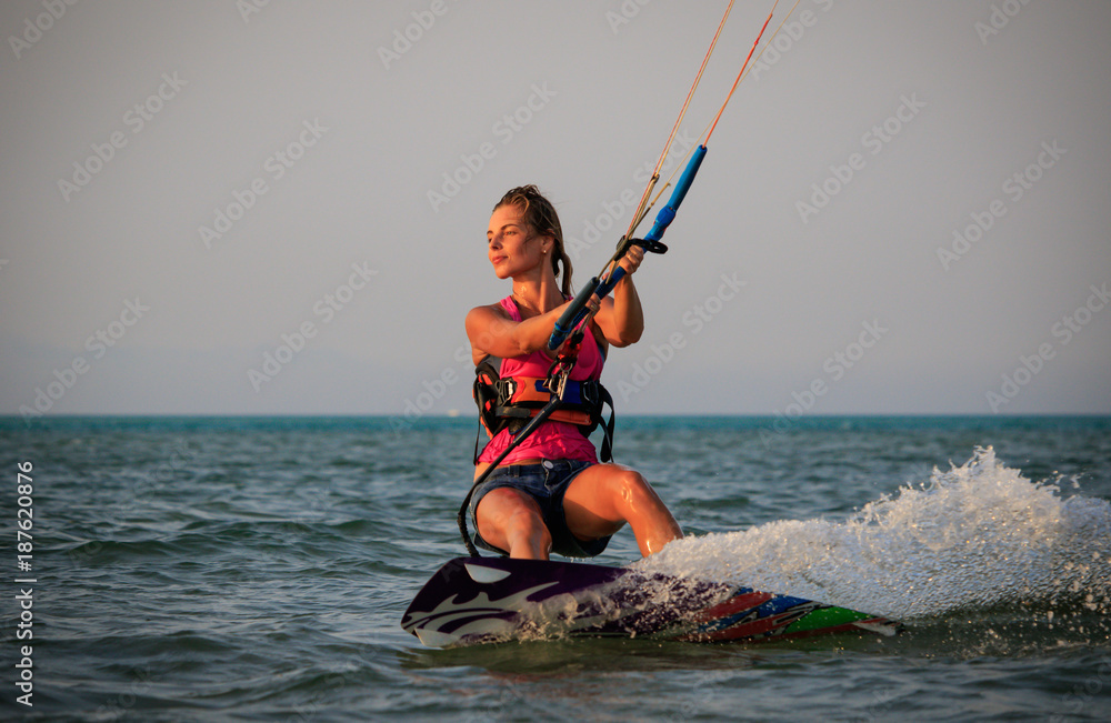 Fototapeta premium Kite surfing girl in sexy swimsuit with kite in sky on kiteboard in the blue sea riding waves saying hi. Recreational activity, water sports, action, hobby and fun in summer time. Kiteboarding sunset