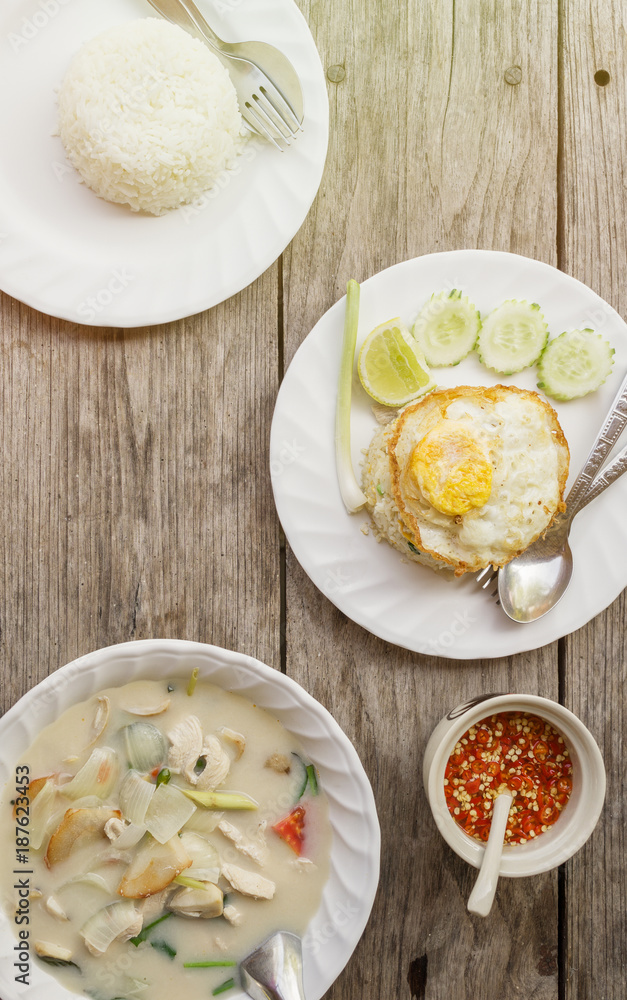 authentic Thai dinner background: Tom kha gai spicy soup, plain rice and pad kra pao with fried egg on top and homemade chilli sauce on wooden table at local cafe. Asian food concept. Text space