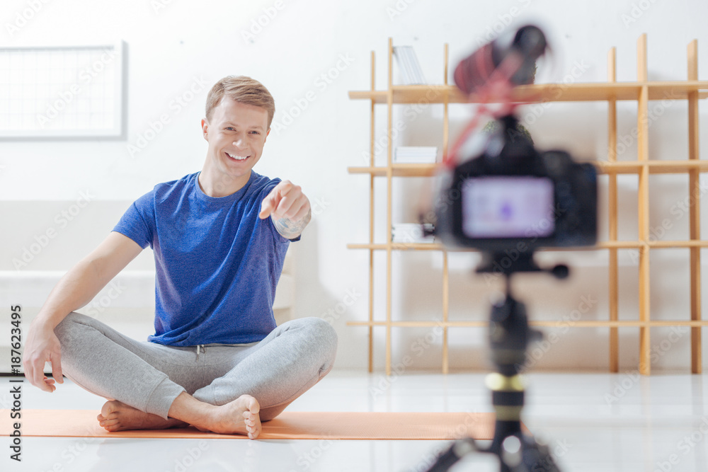 Pointing. Emotional experienced video blogger sitting comfortably on his soft yoga mat and smiling while pointing to the new modern camera in front of him Pointing. Emotional experienced video blogger sitting comfortably on his soft yoga mat and smiling while pointing to the new modern camera in front of him