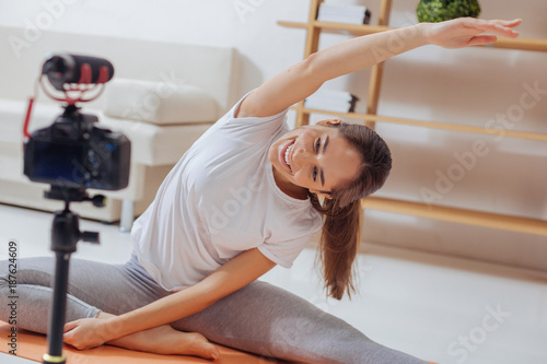 Looking enthusiastic. Emotional cheerful young yoga teacher smiling to the camera while bending on the yoga mat and recording it for her video blog